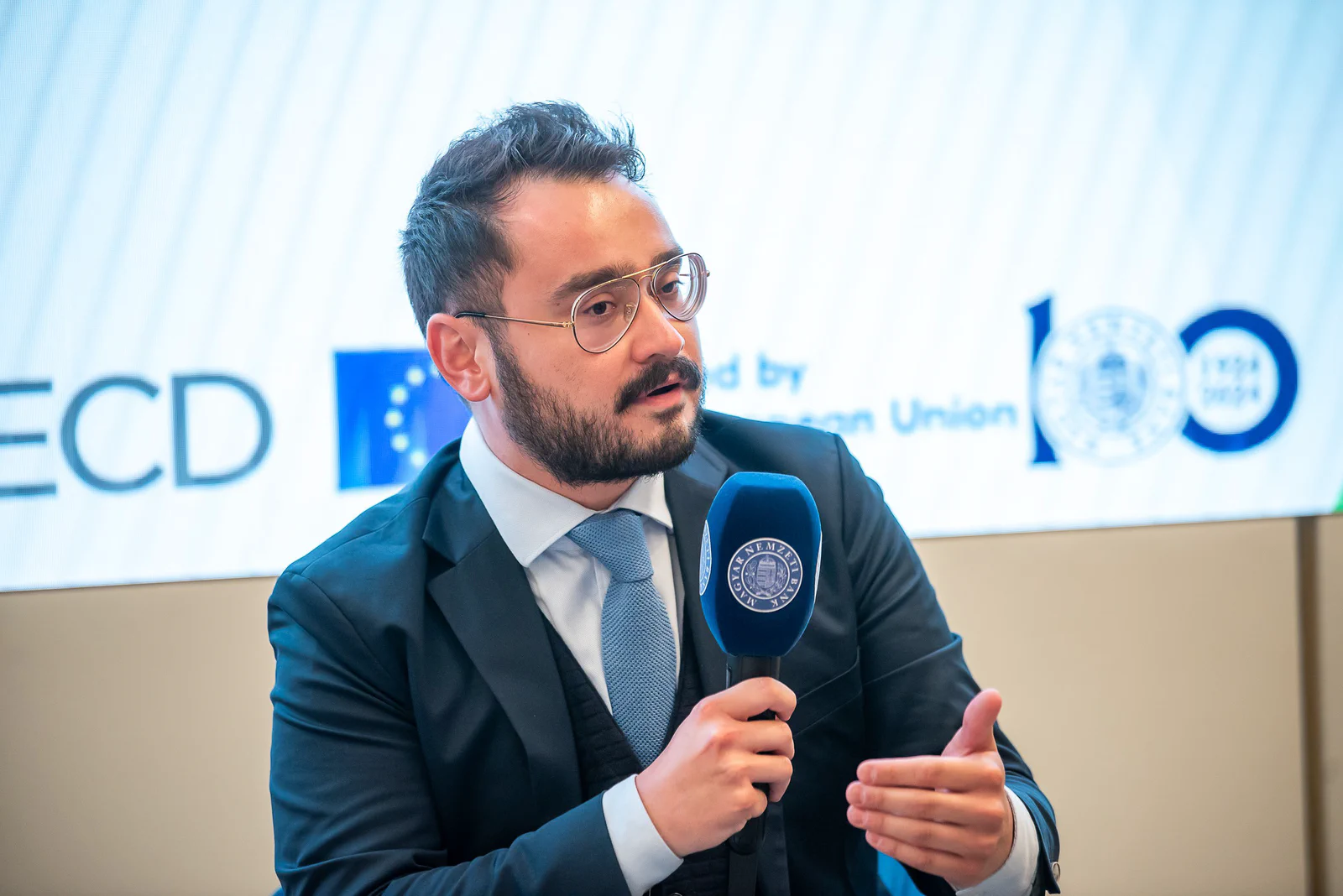 Gürcan Zeren Gülersoy speaking into a Magyar Nemzeti Bank microphone at the OECD–MNB launch event in Budapest.